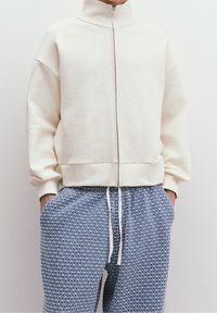 Cremfarbener Zip-Up-Sweatshirt mit hohem Kragen und oversize Passform, kombiniert mit blauen gemusterten Jogginghosen mit Kordelzug und Taschen.
