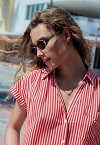 Piros-fehér csíkos rövid ujjú ing, elülső zsebbel. Kerek napszemüveg sötét lencsékkel és türkiz díszítéssel a kereten.