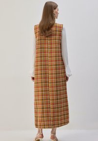 Long gilet à carreaux marron, jaune et rouge, avec une silhouette droite et un design sans manches. Détail de couture au dos visible.