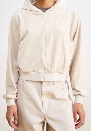 Personne portant un sweat à capuche zippé beige clair avec un petit logo Nike, assorti à un pantalon beige, debout avec les bras détendus le long du corps.