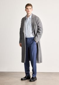 Γκρι μάλλινο παλτό με υφή, σε στυλ πάνω από μπλε και λευκό πουκάμισο με ρίγες, συνδυασμένο με ραμμένο μπλε παντελόνι και μαύρα παπούτσια.