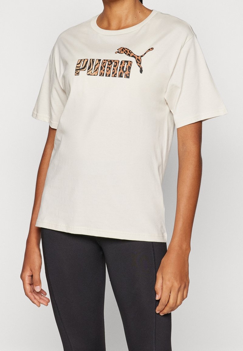 Personne portant un t-shirt blanc Puma avec un logo imprimé léopard associé à un legging noir ajusté sur un fond uni.