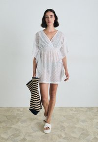 White lace cover-up with a V-neck, short sleeves, and flared design. The model holds a black and beige striped tote. White slip-on sandals.