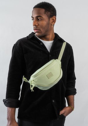 Marsupio in nylon verde chiaro con una texture liscia e chiusura con zip, indossato attraversato sul corpo. Presenta una tracolla regolabile e un dettaglio del logo.