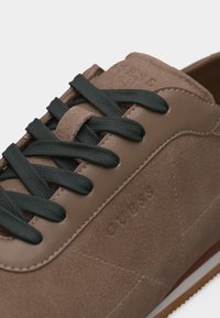 Καφέ σουέντ αθλητικό παπούτσι με μαύρα κορδόνια, με τη σήμανση "GUESS" ανάγλυφη στο πλάι και λεπτομερείς ραφές στις άκρες.