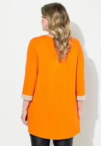 Orange Tunika mit Dreiviertelärmeln. Verfügt über Häkelakzente an den Ärmeln, glattes Material und eine lockere Passform, getragen über schwarzen Hosen.