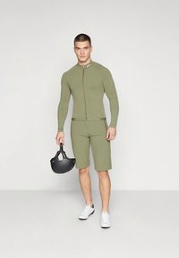 Olivengrønt treningsantrekk med lange ermer og shorts, med glidelås foran. Holder en svart hjelm, kombinert med hvite joggesko.