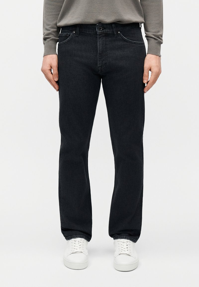 Άντρας που φοράει μαύρο τζιν με ίσια γραμμή, λευκά αθλητικά παπούτσια και γκρι μακρυμάνικη μπλούζα, στέκεται μπροστά σε λευκό απλό φόντο.