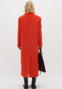 Röd oversized jacka med en texturerad finish, som har en slits bak, långa ärmar och en nedvikt krage, tillsammans med en svart väska.