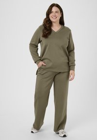 Femme debout et souriante, portant un haut V à manches longues vert olive, un pantalon assorti et des baskets beiges sur fond blanc.