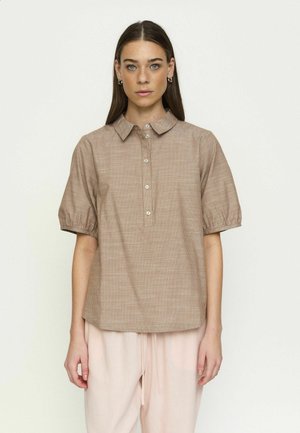 Žena s dugom smeđom kosom nosi bež košulju s kratkim rukavima i gumbima te svijetlosive široke hlače, stojeći na jednostavnoj svijetloj pozadini.