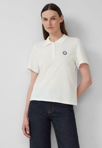 Weißes Polo-Shirt mit klassischem Kragen, Knopfleiste und einem runden blauen Patch auf der Brust. Kurze Ärmel und lockere Passform.