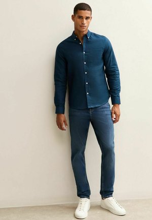 Donkerblauwe overhemd met knopen en een borstzak, gecombineerd met lichtblauwe jeans en witte sneakers. Glad materiaal en een getailleerd ontwerp.