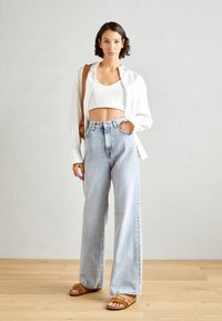 Lichtblauwe hooggewaadde jeans met wijde benen, gecombineerd met een witte cropped top en een witte overhemd. Bruine sandaaltjes met bandjes maken de look af.