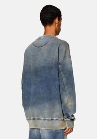 Felpa in denim blu sfumato, con colletto a girocollo, spalle scese e una finitura testurizzata. I bordi mostrano un leggero sfilacciamento.