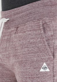 Bordeauxfarbene, strukturierte Jogginghosen mit einem Kordelzugbund, seitlichen Taschen und einem weißen, dreieckigen Logopatch am linken Bein.