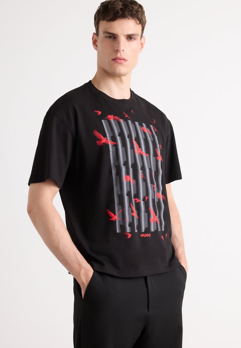 T-shirt noir avec un design graphique comprenant des oiseaux rouges et des rayures verticales grises. Le tissu est en coton avec des manches courtes et un col rond.