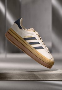 White sneaker with tan and navy accents, featuring three dark blue stripes, textured suede, and a layered rubber sole. Brass "Gazelle" logo.