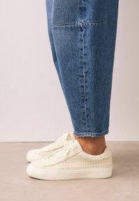 Baskets tissées blanches avec une surface texturée, bout rond et semelle en caoutchouc blanc plat, associées à un pantalon en jean bleu.