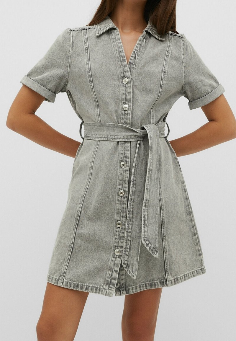 Femme portant une robe en jean grise à manches courtes, boutonnée devant avec col et ceinture nouée à la taille, debout les mains croisées dans le dos.