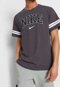 Šedé bavlněné tričko s nápisem "NIKE" v tučném písmu, s bílými proužky na rukávech a malým logem Nike pod textem.
