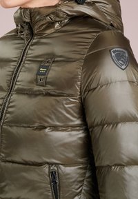 Primer plano de una chaqueta acolchada color verde oliva con cremallera, parches en el pecho y en las mangas con el logo "Blauer U.S.A.".