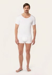 Witte aangesloten short-sleeve shirt met V-hals, gecombineerd met witte boxershorts, getoond op een mannelijke model, blootsvoets tegen een neutrale achtergrond.