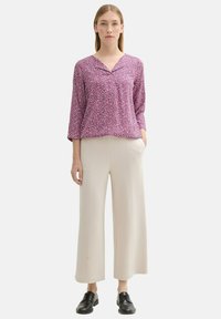 Lila und rosa gemusterte Bluse mit V-Ausschnitt und Dreiviertelärmeln, kombiniert mit beigen weiten Hosen und schwarzen Schuhen.