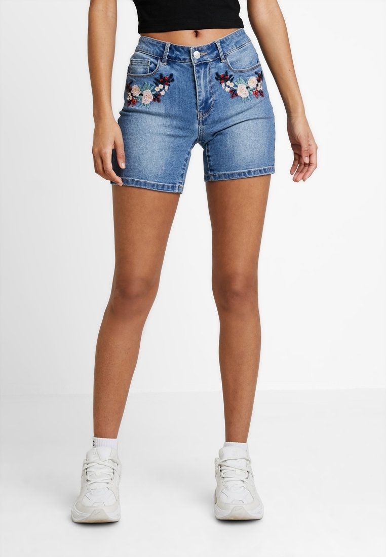 Mujer con pantalones cortos de mezclilla azul con bordados florales en los bolsillos delanteros, zapatillas blancas y un top negro corto, de pie contra un fondo blanco.