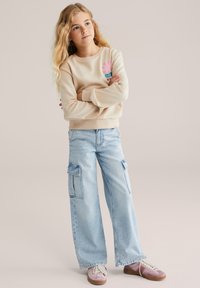 Jauna meitene ar gariem blondiem matiem stāv ar saliktām rokām, ģērbusies bežā džemperī ar rozā dizainu, gaismas zilos cargo džinsos un rozā sporta apavos.