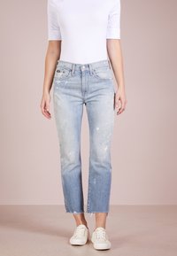 Světle modré, opotřebované džíny s barvovými skvrnami, rovné nohavice, neupravené lemy a vysoký pas, doplněné bílými teniskami.