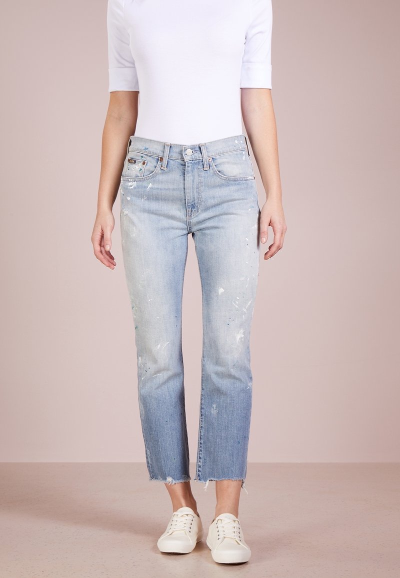 Světle modré, opotřebované džíny s barvovými skvrnami, rovné nohavice, neupravené lemy a vysoký pas, doplněné bílými teniskami.