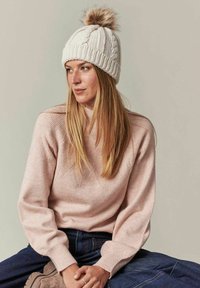 Geweven crèmekleurige beanie met een pompon bovenop, gecombineerd met een lichtroze trui met een gestructureerd honingraatpatroon. Donkerdenimbroek.