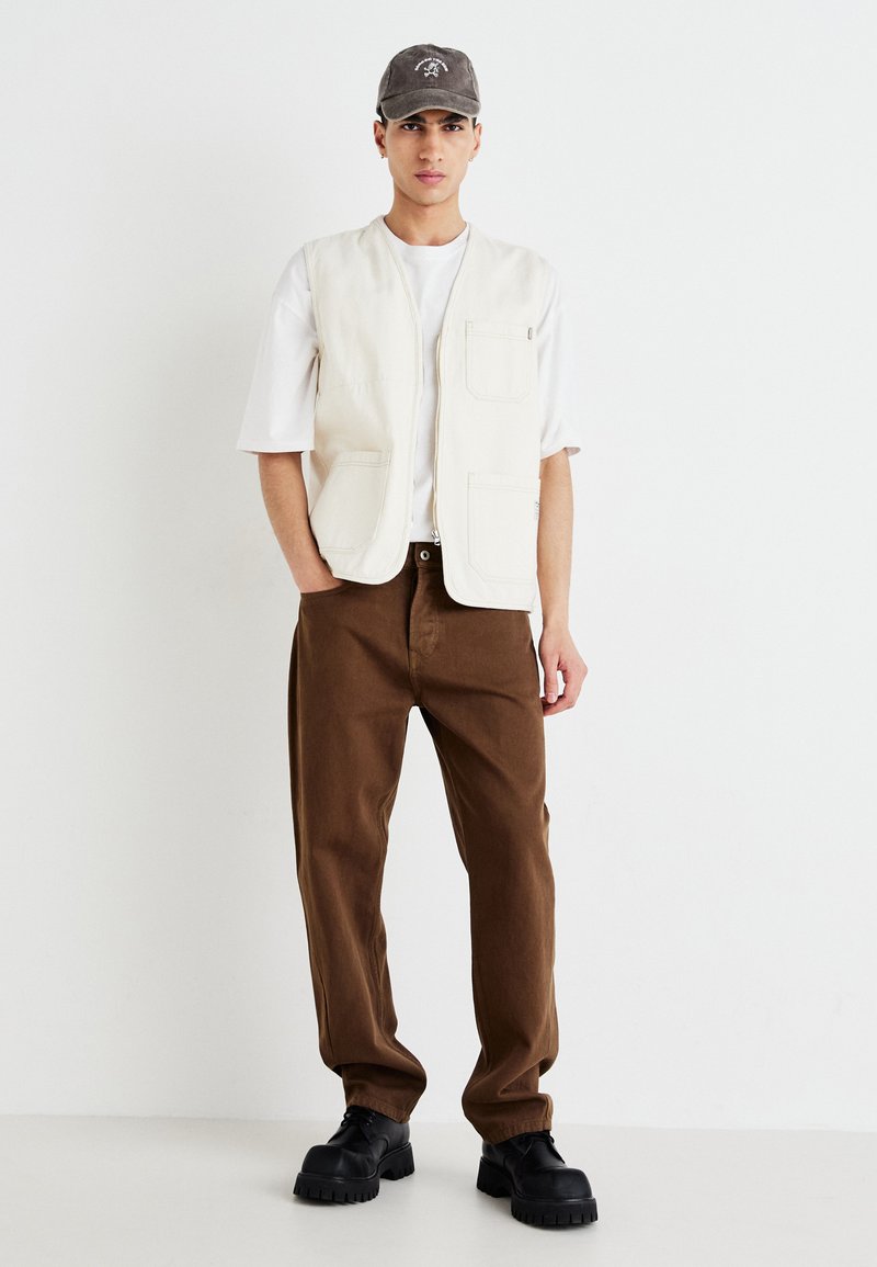 Beige mouwloze vest met voorzakken, gedragen over een wit t-shirt. Bruine broek met wijde pijpen en zwarte chunky schoenen maken de look compleet. Grijze pet toegevoegd.