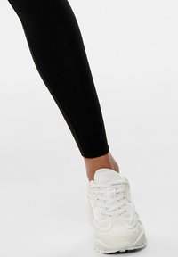 Leggings noirs ajustés avec une texture lisse, associés à des baskets de sport blanches avec des lacets et une semelle texturée.