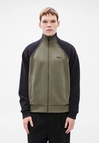 Cipzáros dzseki, mely olívazöld testből és fekete ujjú részből áll, fehér csíkozással és első zsebekkel. A mellkason logó látható.