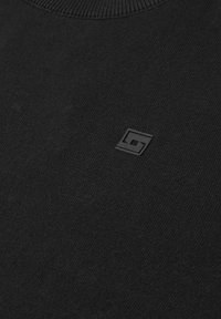 Tissu tricoté noir avec un petit logo géométrique noir en relief près de la partie supérieure de la poitrine sous le col.