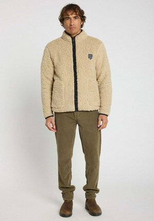 Homme portant une veste polaire beige, un pantalon en velours côtelé olive et des bottes marron, debout devant un fond blanc.