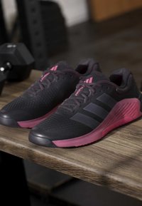 Ζευγάρι μαύρα και ροζ παπούτσια τρεξίματος Adidas τοποθετημένα σε ξύλινη επιφάνεια με μαύρο αλτήρα στο φόντο.