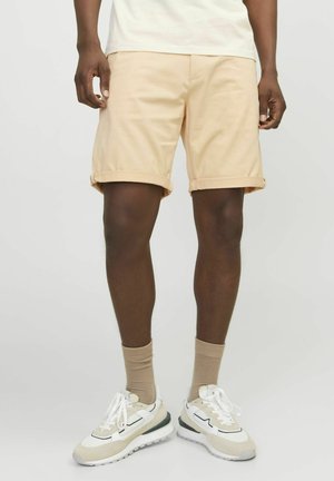 Pantalones cortos de algodón beige claro con puños enrollados, que cuentan con un frente plano y dos bolsillos laterales, combinados con calcetines neutros y zapatillas deportivas.