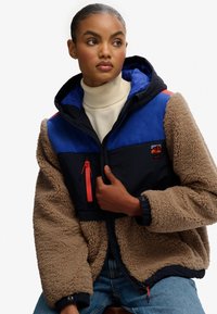 Fleece-gevoerde jas met een marineblauw en blauwe kleurblokontwerp, oranje ritsaccenten en een capuchon. Bevat een ritsborstzak.