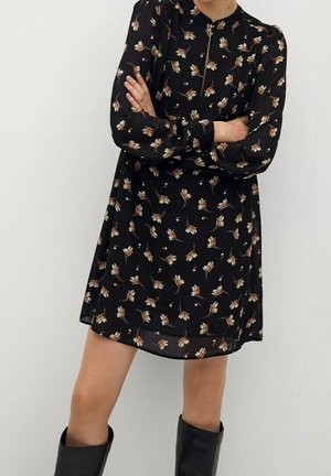 Frau, die ein schwarzes, mit Blumen bedrucktes Langarmkleid mit einem Reißverschluss-Ausschnitt und schwarzen kniehohen Stiefeln trägt und mit verschränkten Armen dasteht.