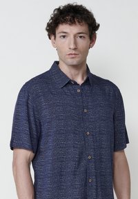 Camicia button-up a maniche corte di colore blu navy con un motivo testurizzato, caratterizzata da fili chiari a contrasto e bottoni in legno.