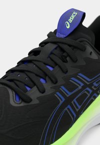 Zapatilla deportiva negra con acentos azules y parte superior texturizada. Cuenta con un sistema de cierre con cordones, lengüeta acolchada y suela de un verde neón contrastante.