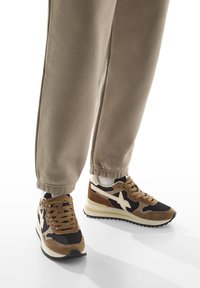 Des baskets en suède marron et noir avec un logo étoile crème, portées avec un pantalon de jogging beige et des chaussettes blanches sur fond blanc.