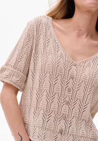 Beżowy sweter na ramiączkach z dekoltem w kształcie litery V, krótkimi podwiniętymi rękawami i geometrycznym wzorem. Z przodu zdobią go sześć ozdobnych guzików.