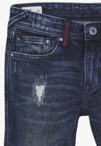 Slim fit tmavě modré denimové džíny s vybledlými detaily, roztrhanými koleny, předními kapsami a červeným štítkem značky uvnitř pásku.