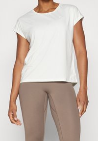 Camiseta deportiva blanca de manga corta con escote redondo y textura suave, combinada con leggings marrones ajustados que presentan detalles de costuras.