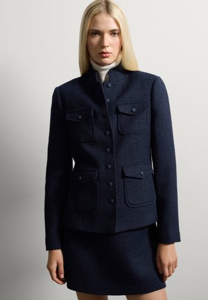 Chaqueta de tweed navy con cuello alto, doble bolsillo en el pecho y cierre de botones, combinada con una falda a juego. Textura suave, diseño ajustado.
