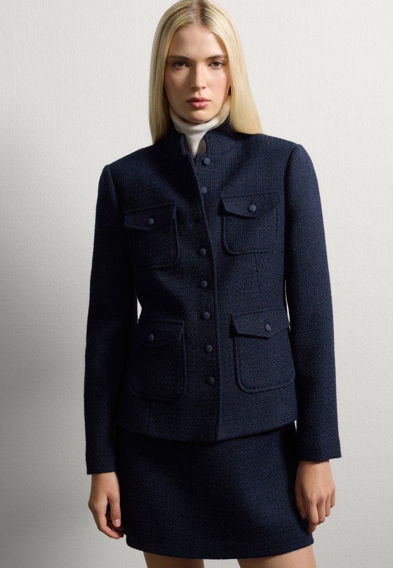 Giacca in tweed navy con colletto alto, due tasche sul petto e chiusura a bottone, abbinata a una gonna coordinata. Tessuto liscio, design sinuoso.